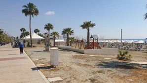 A beachside promenade with palm trees, a covered seating area, and a row of sun loungers. - Marina's Rooms (Larnaca)