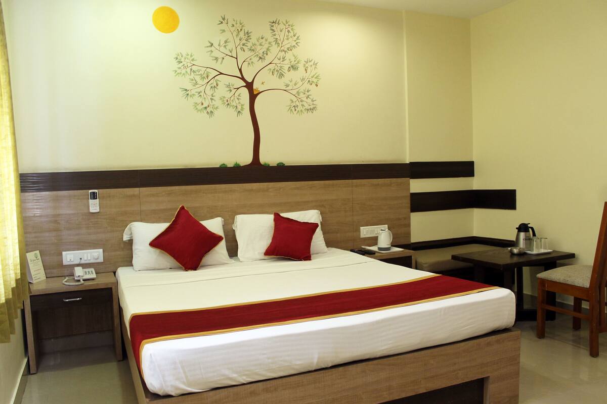 A hotel room with a bed, two red pillows, a nightstand with a phone, a table with a kettle, and a wall decoration of a tree.
