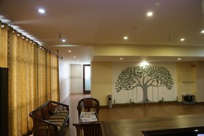 Un vestíbulo de hotel con un mostrador de recepción de madera, dos sillas de madera, un mural grande con motivo de un árbol en la pared y una cortina con estampado ondulado.