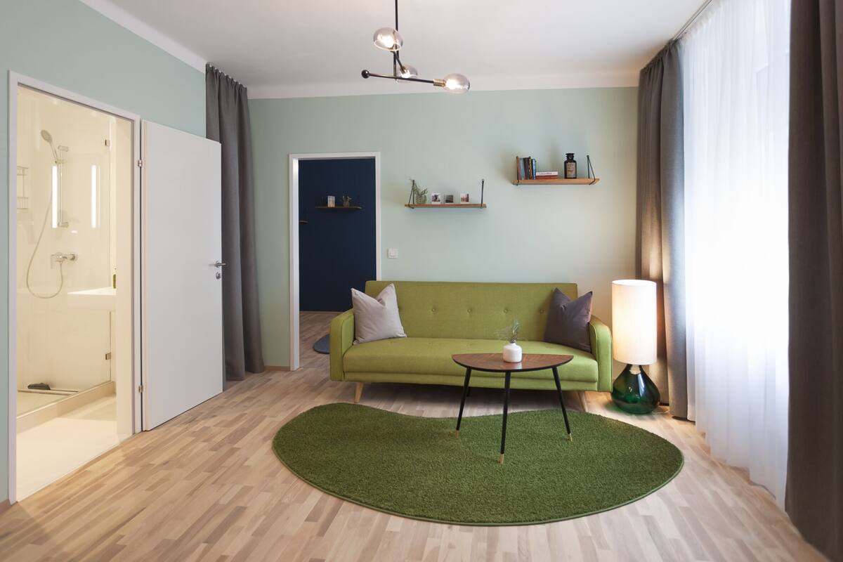 Ett modernt vardagsrum med en grön soffa, ett litet runt bord och en grön matta. Till vänster finns en duschhörna och en dörr som leder till ett annat rum.