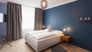 Quarto moderno com cama grande, mesa de cabeceira com luminárias, uma cadeira e vista da cidade através de cortinas finas.