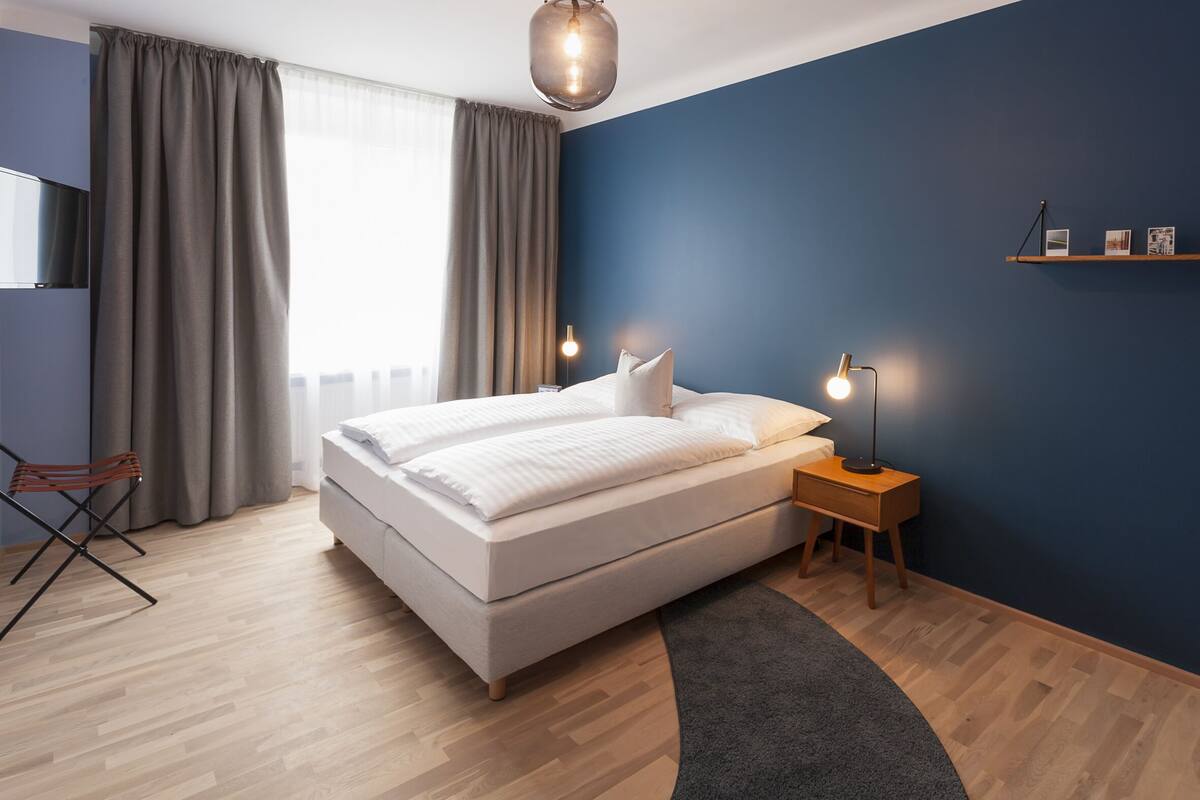 Ett modernt sovrum med en stor säng, ett sängbord med lampor, en stol och utsikt över staden genom tunna, genomskinliga gardiner.