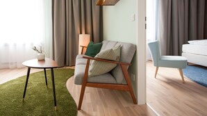 Uma sala de estar moderna com uma poltrona cinza, uma mesa redonda pequena com uma planta, e outra poltrona azul. Há um sofá branco ao fundo e um abajur sobre uma mesinha lateral.