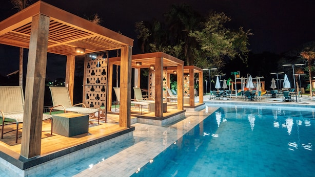 Zona de piscina nocturna con pérgolas de madera, sillones de descanso y sombrillas.