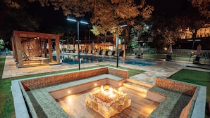 Un área de piscina exterior de noche con una fogata, sillas de descanso y una zona de estar cubierta.