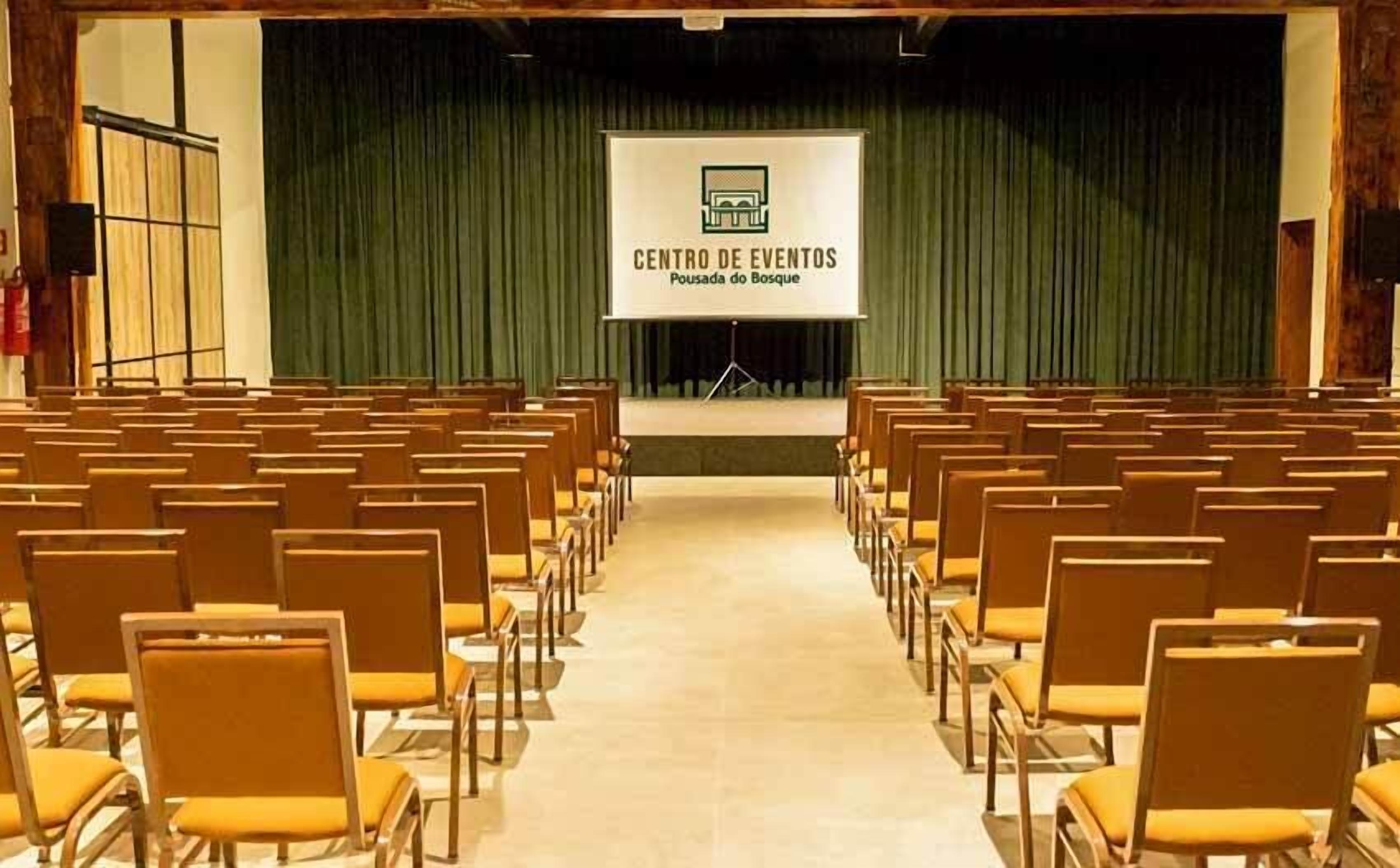Un auditorio con hileras de sillas amarillas, un escenario con una pantalla que muestra 