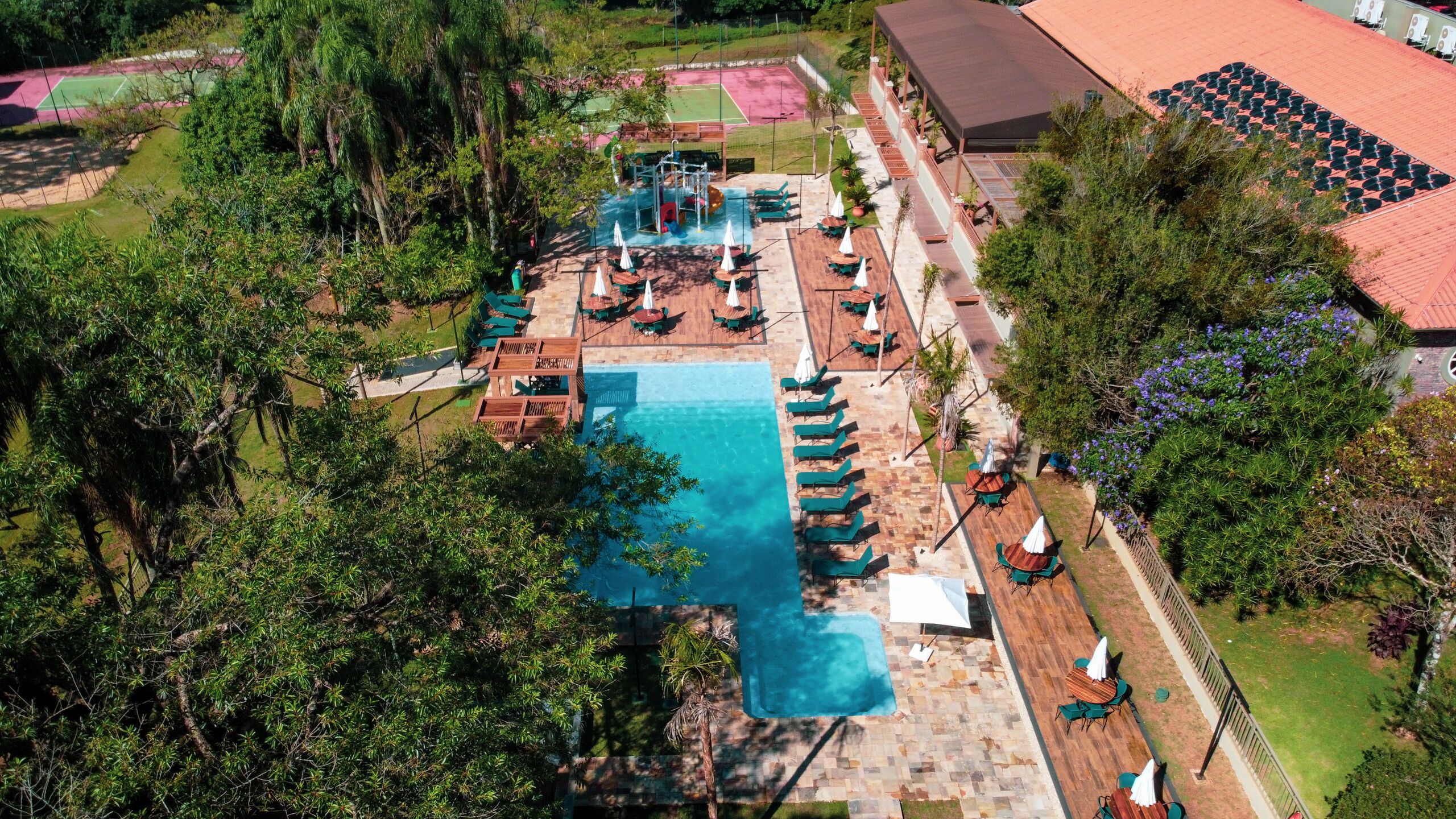 An aerial view of a resort with a swimming pool, sun loungers, and a tennis court.