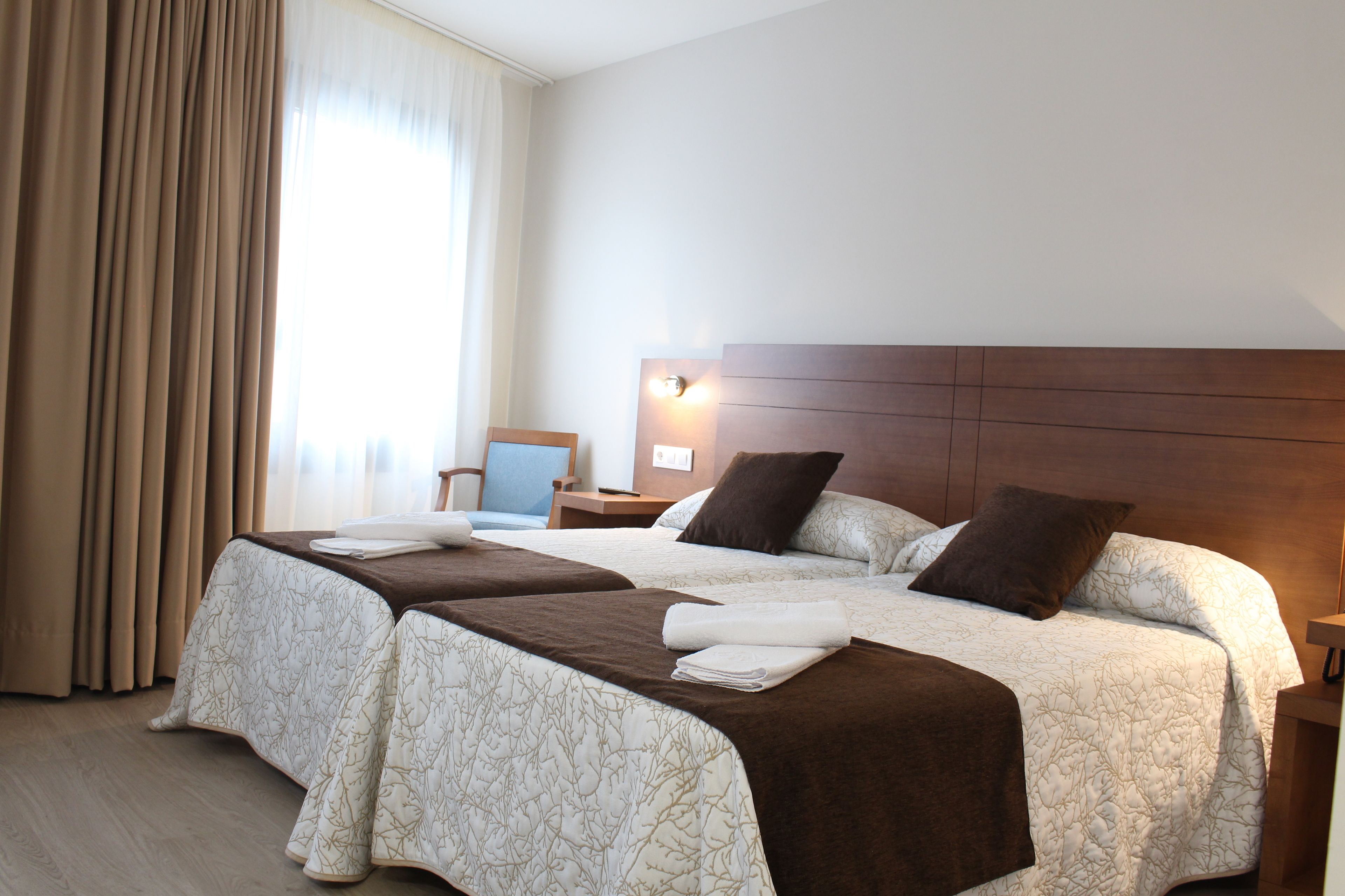 Habitación de hotel con dos camas, cabecero de madera, mesita de noche y una silla.