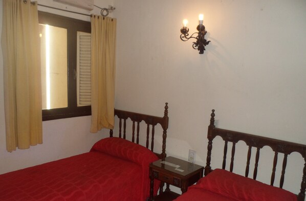 A room with two wooden beds, a small table, a wall-mounted light fixture, and a window with curtains. - Emperatriz Hotel (Santa Fe)
