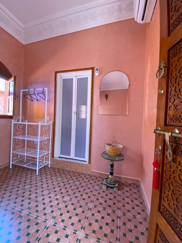 Una habitación con piso estampado, una puerta blanca con panel de vidrio, un estante metálico, una mesita con una cesta y un espejo decorativo.