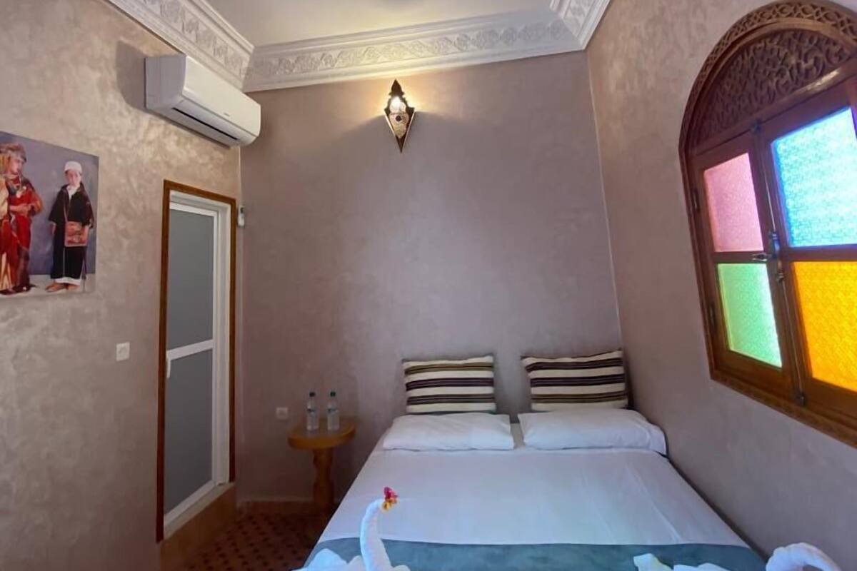 A hotel room with a bed, two swan-shaped towels, a wall-mounted air conditioner, a decorative light fixture, a framed picture, and a window with a colorful view.