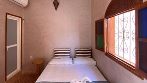 A hotel room with a bed, a small bedside table, a wall-mounted air conditioner, a decorative wall piece, and a window with decorative iron bars. - Dar Dikrayat (Marrakech)