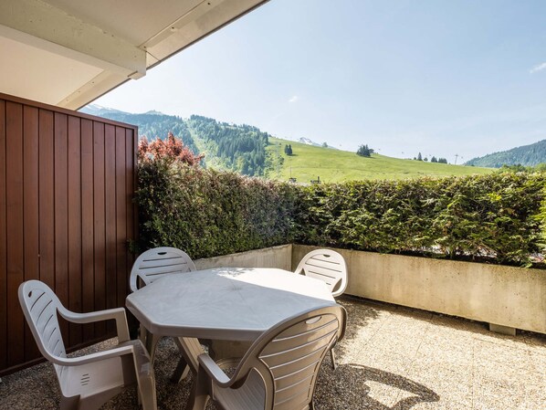 A patio with a table and chairs, a wooden wall, and a view of a green hillside.