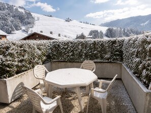 A snow-covered outdoor seating area with a table and chairs, surrounded by snow-laden bushes and a snowy mountain backdrop.