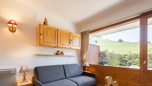 A room with a sofa, wooden cabinets, a small table, and a window with a view of a green landscape. - Renovated 3* studio in La Clusaz with south terrace (La Clusaz)