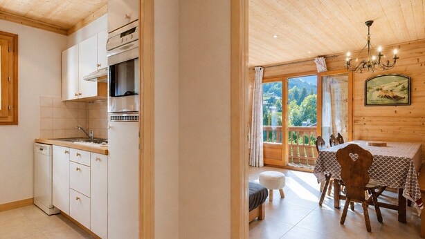 Une cuisine avec des meubles blancs, une cuisinière et un lave-vaisselle. On y trouve un coin repas avec une table et des chaises, et une vue sur l’extérieur grâce à une grande fenêtre.
