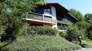 A two-story building with a balcony, surrounded by dense greenery. - Spacious 3-bedroom apartment near slopes, pets allowed, parking (La Clusaz)