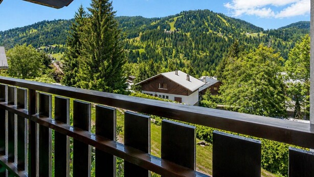 A balcony with a view of a mountainous landscape, a wooden house, and a forested area. - Spacious 3-bedroom apartment near slopes, pets allowed, parking (La Clusaz)