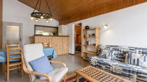 A cozy living room with a wooden ceiling, a sofa, a chair, a coffee table, and a kitchen area in the background. - Spacious 3-bedroom apartment near slopes, pets allowed, parking (La Clusaz)