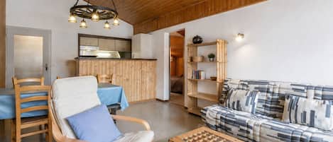 A cozy living room with a wooden ceiling, a sofa, a chair, a coffee table, and a kitchen area in the background.