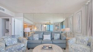 A living room with a grey sofa, two patterned armchairs, a round coffee table, and a large mirror reflecting the room. - Luxurious Beachfront Corner Unit at Seaside Beach & Racquet 4407 (Orange Beach)