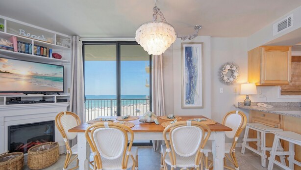 A dining area with a wooden table and wicker chairs, a television on the wall, and a view of the beach through the window. - Luxurious Beachfront Corner Unit at Seaside Beach & Racquet 4407 (Orange Beach)