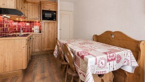 Een kleine keuken met houten kastjes, een magnetron en een gootsteen. Er is een tafel met een tafelkleed met een patroon en een bed met een houten hoofdbord.