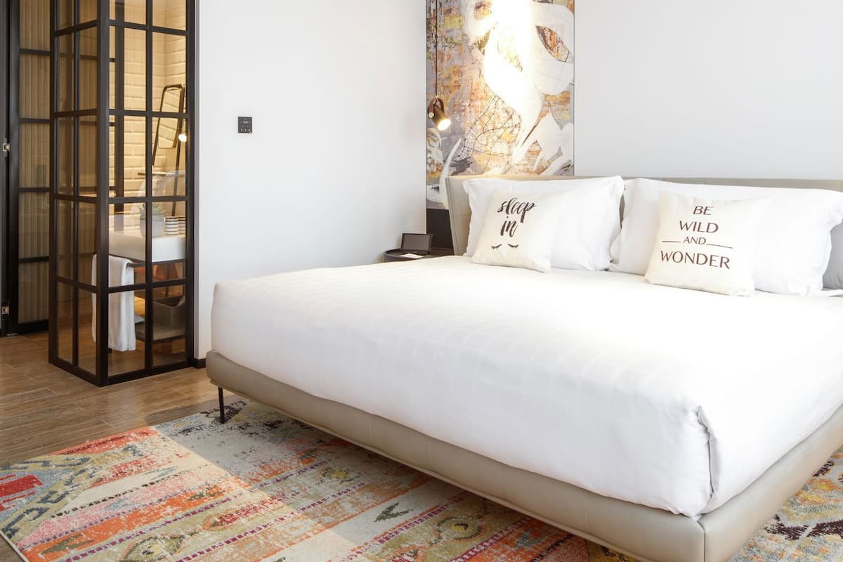 Un dormitorio moderno con una cama grande, ropa de cama blanca, una alfombra colorida, una mesita de noche con una lámpara y una pared decorada con un mapa.