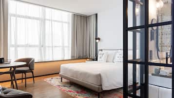 A modern hotel room with a large bed, a desk with chairs, and a view of the city through large windows.