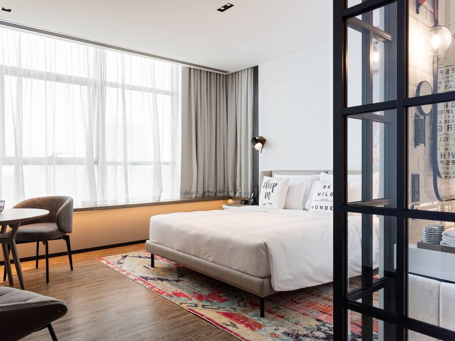 A modern hotel room with a large bed, a desk with chairs, and a view of the city through large windows.