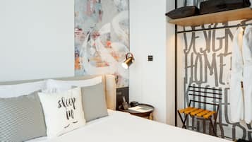 Una camera d'albergo moderna con un letto, cuscini, un comodino, una lampada, una sedia e un grande quadro astratto appeso al muro.
