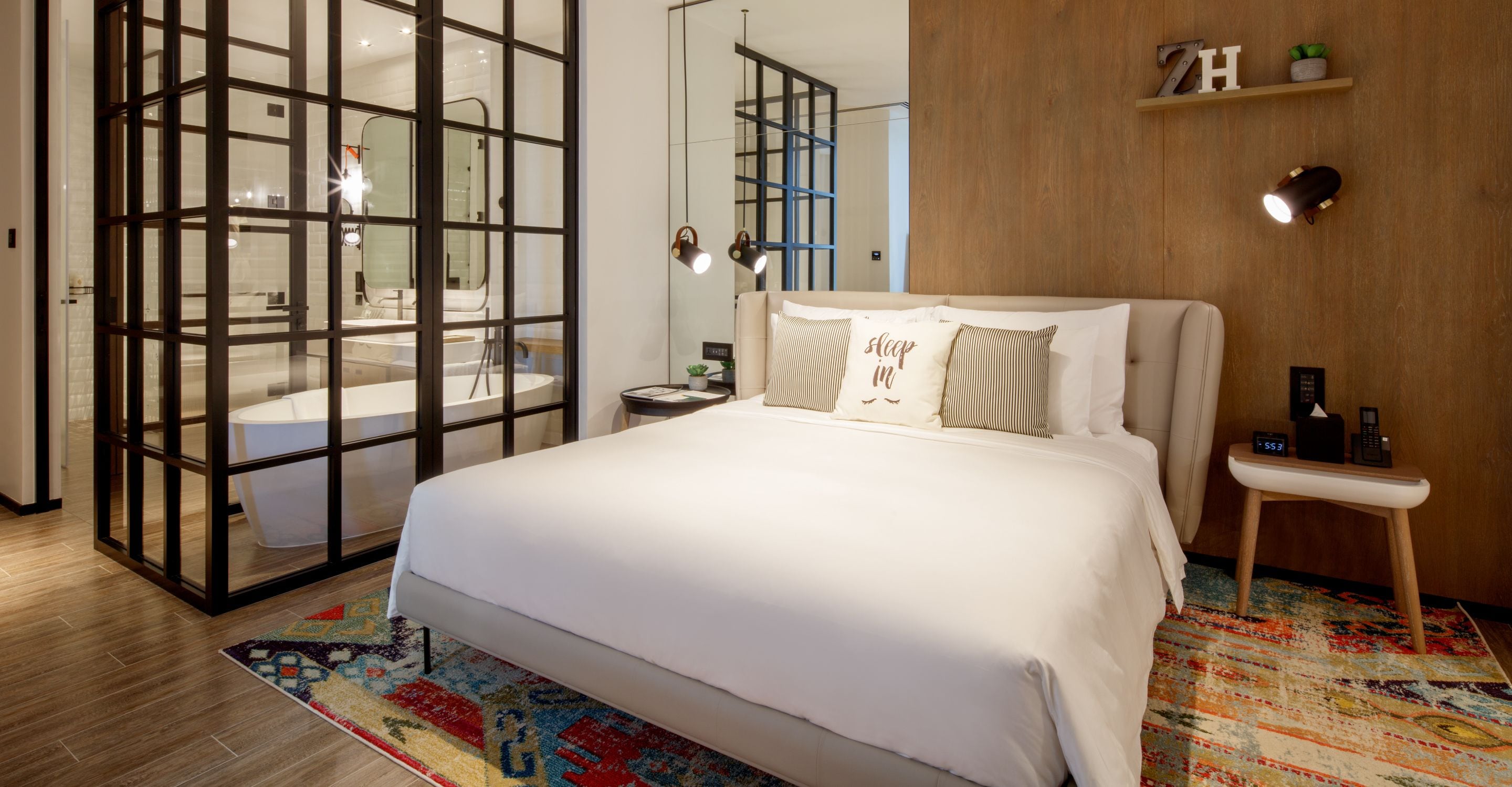 Une chambre d’hôtel moderne avec un grand lit, un plancher en bois, un tapis coloré, une table de chevet avec une lampe et une petite étagère ornée d’objets décoratifs.