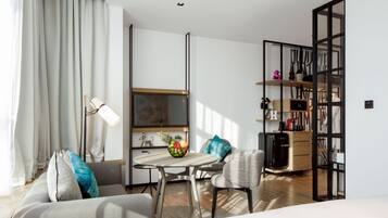 A modern hotel room with a sofa, a dining table, a small kitchen area, and a separate seating area with a chair.