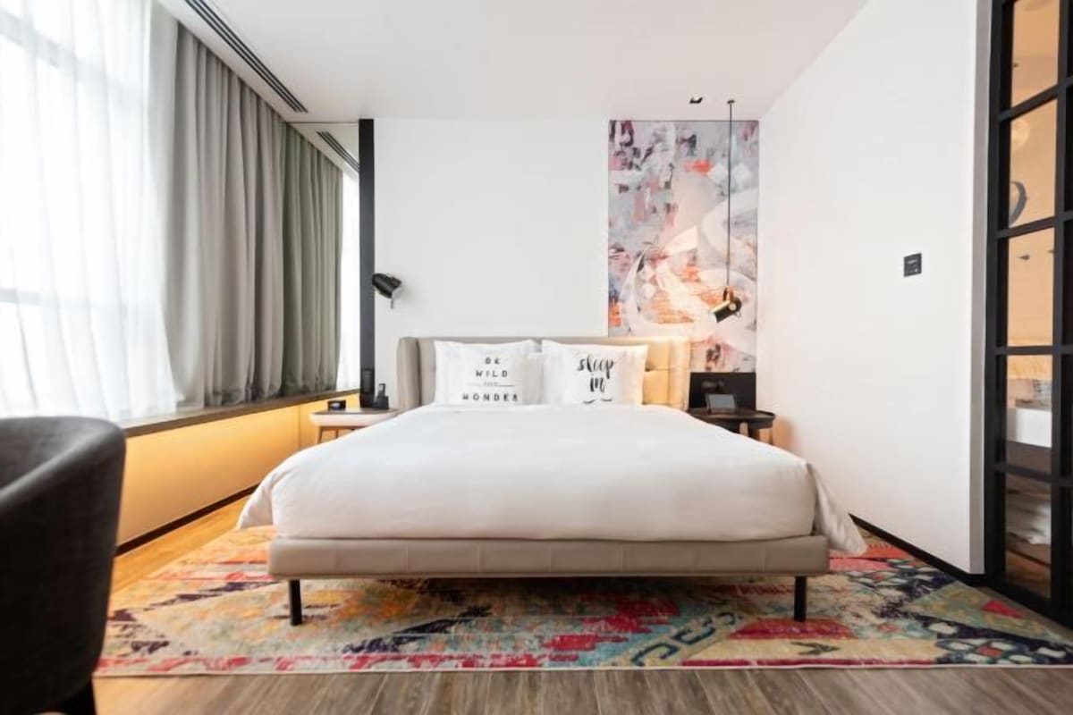 Een hotelkamer met een bed, een nachtkastje, een lamp en een groot abstract schilderij aan de muur.