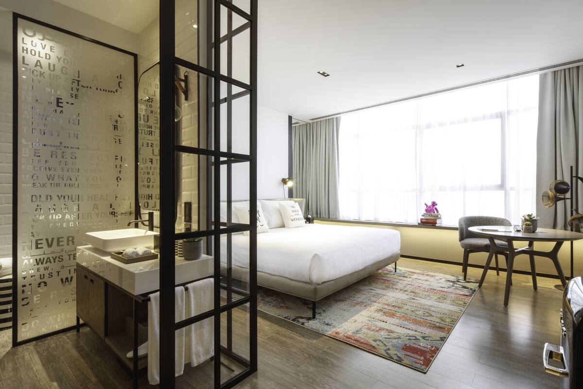 Una habitación de hotel moderna con una cama grande, una mesita de noche, un escritorio pequeño, una silla, una ventana con cortinas y un baño con lavamanos y espejo.
