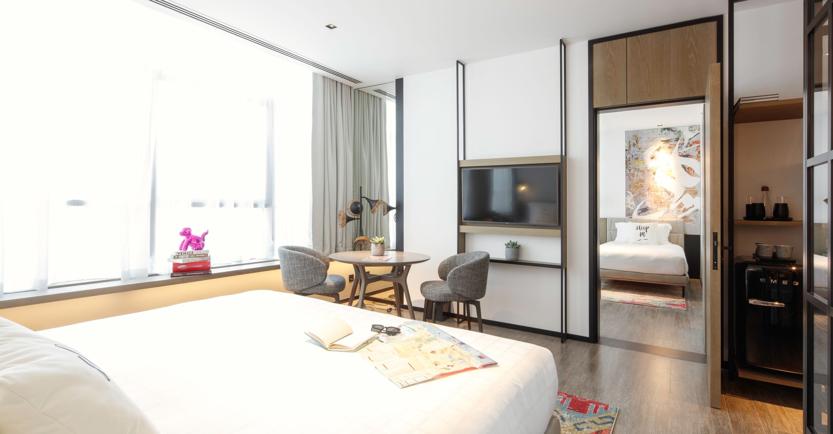 Habitación de hotel con cama, escritorio con dos sillas, televisor, minibar y un ventanal grande con cortinas.