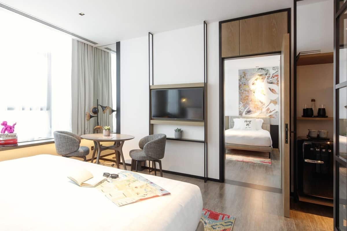 Habitación de hotel con cama, escritorio, dos sillas, televisor, minibar y un ventanal grande.