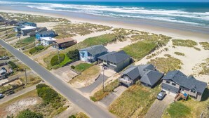 Zona residencial costera con casas junto a una playa, con una carretera y dunas cubiertas de hierba.