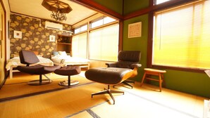 A traditional Japanese-style room with tatami flooring, a low table, two cushioned seating areas, a bed with a wooden headboard, and large windows with blinds. - Super Tomato House Takayama (Takayama)