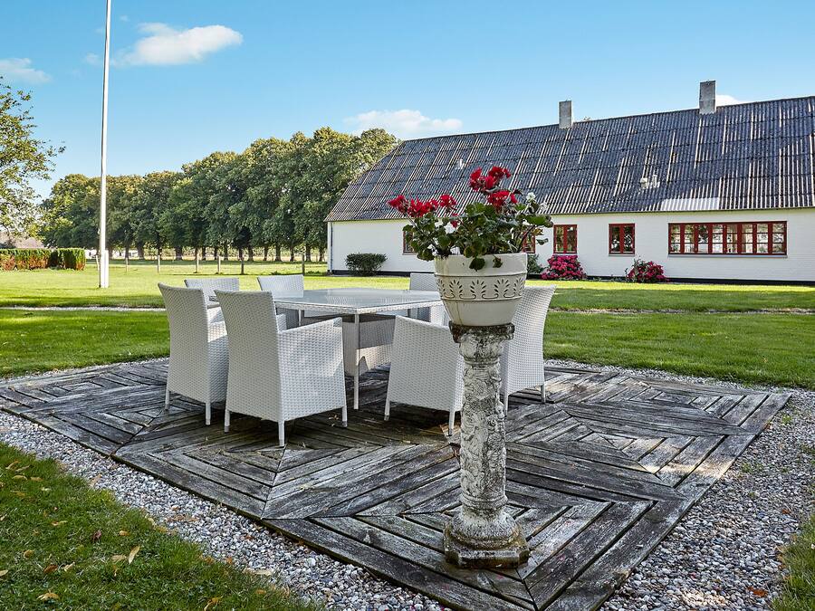 Un pati amb una taula i cadires, una jardiniera decorativa plena de flors, i una casa blanca al fons.