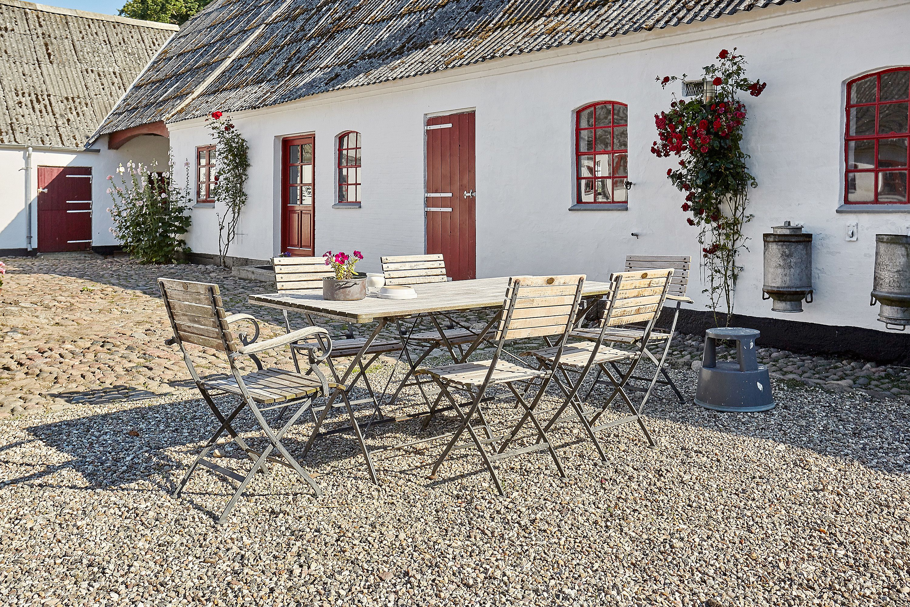 A white building with red doors and windows, a gravel courtyard, a wooden table with chairs, and a rose bush.