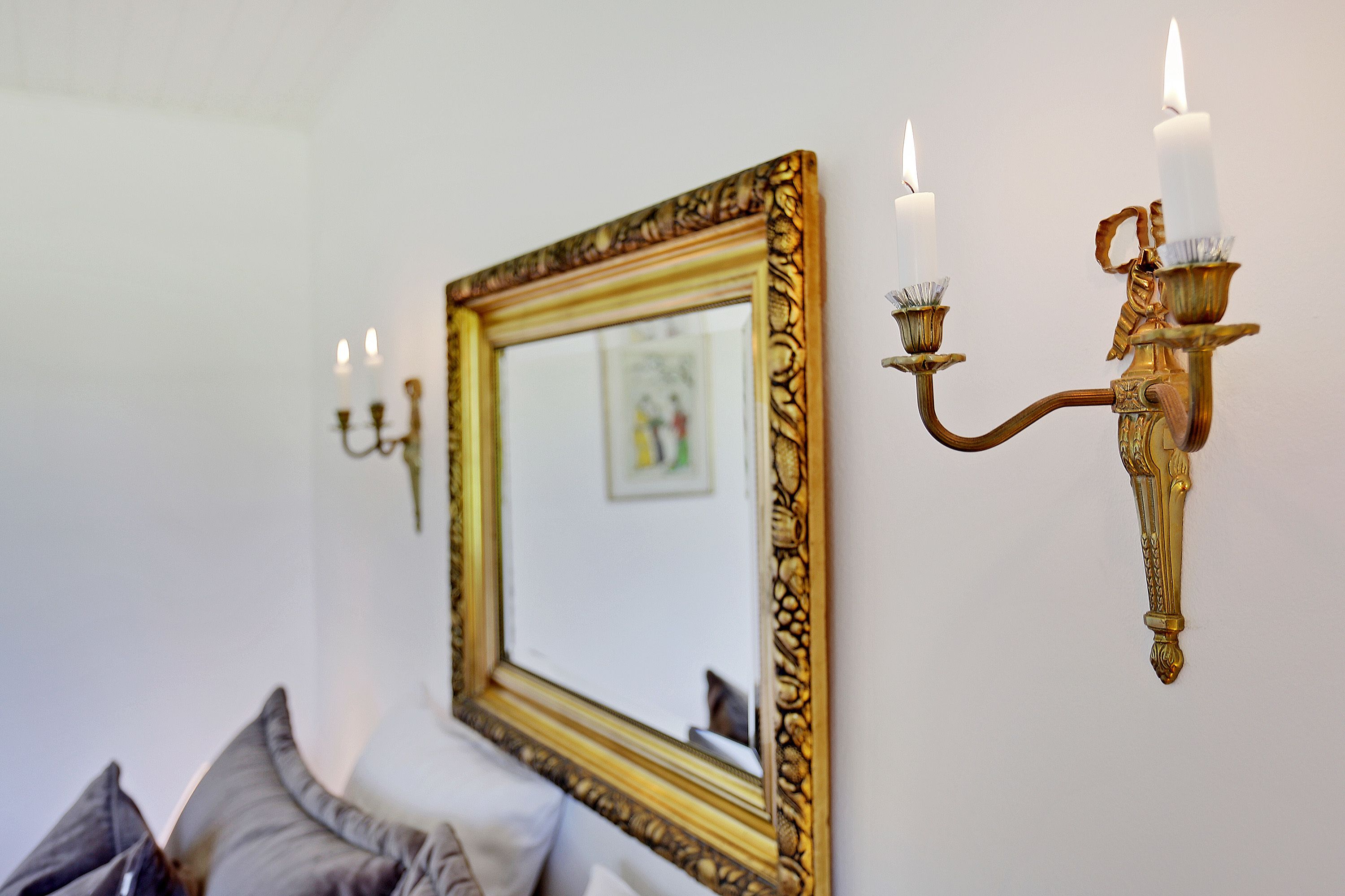 Una habitación con un espejo grande y ornamentado, un candelabro fijado a la pared con velas encendidas, y una cama con almohadas.