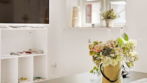 A living room with a TV on a stand, a bookshelf, a window with a plant, and a vase with flowers on a table. - Bed and Breakfast Åløkke (Nyborg)
