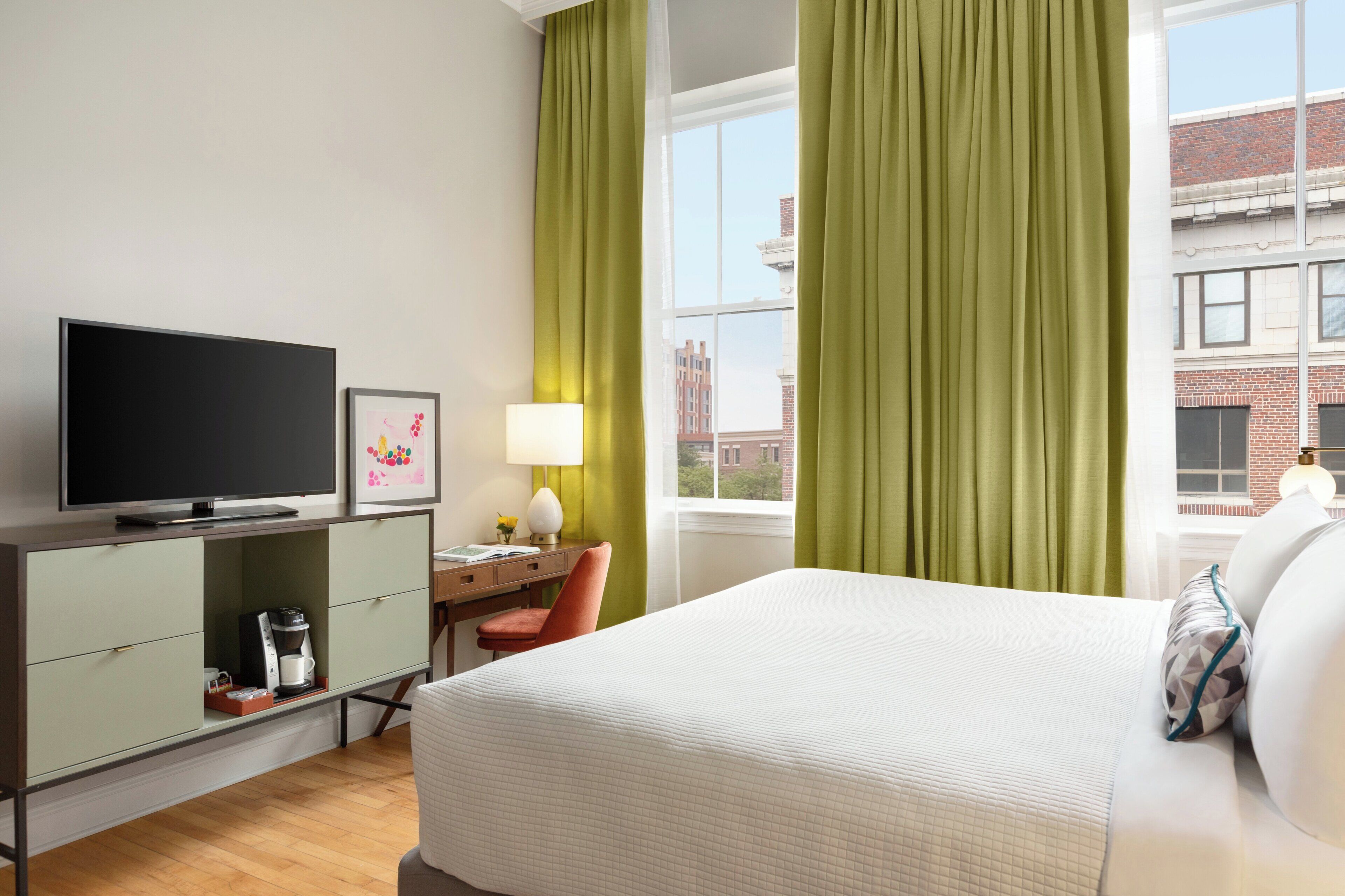 Une chambre d’hôtel comprenant un lit, un bureau avec une télévision, une chaise, une lampe et une fenêtre avec des rideaux.