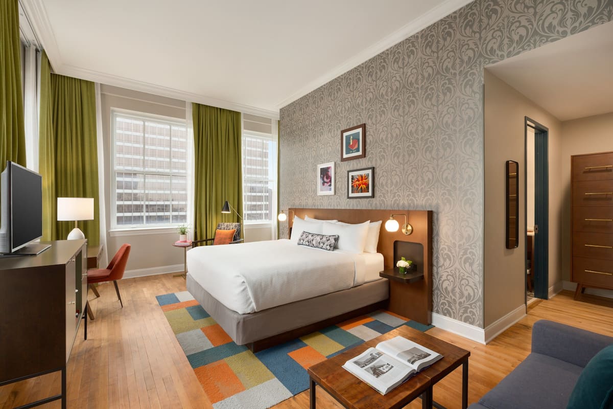 A hotel room with a bed, a desk, a chair, a sofa, and a small table. There is a window with curtains, a wall with framed pictures, and a door.