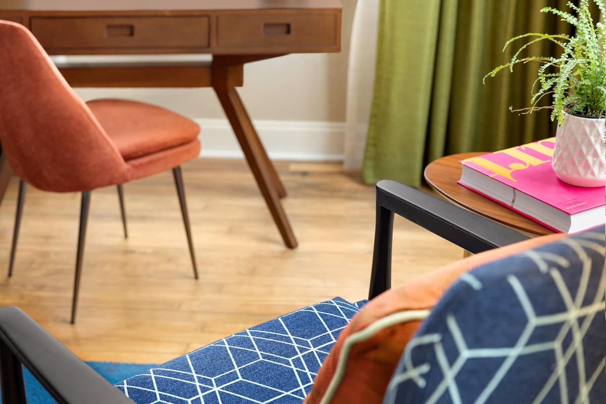 A room with a wooden desk, a pink chair, a blue patterned cushion, a small potted plant, and a book on a side table.