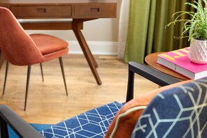 Una habitación con un escritorio de madera, una silla rosa, un cojín azul estampado, una planta pequeña en maceta y un libro sobre una mesita.