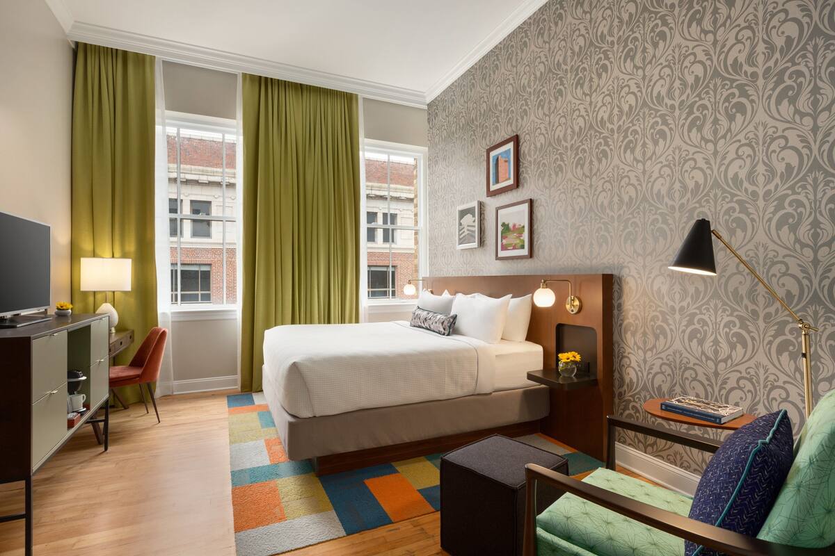 A hotel room with a bed, a desk, a chair, a lamp, and a view of the city.