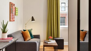 A modern living room with a grey sofa, a small table with a book and a vase of flowers, a floor lamp, and a view of a brick building through a window with green curtains. - Hotel Indigo Winston-Salem Downtown by IHG (Winston-Salem)
