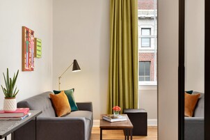 Una sala moderna con un sofá gris, una mesita con un libro y un jarrón de flores, una lámpara de pie y vista a un edificio de ladrillo a través de una ventana con cortinas verdes.